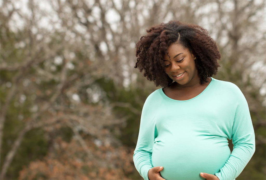 Black pregnant woman in mint-green shirt holding her belly.